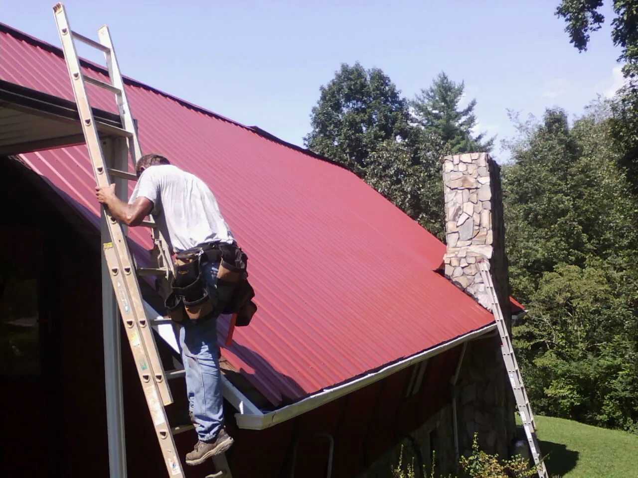 Roofer on a ladder beside a red metal roof and stone chimney — Soffit & Fascia Repair by Roofing Brothers in Church Creek